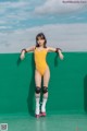 A woman in a yellow bodysuit on roller skates leaning against a green wall.